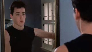 John Cusack in a black tank top standing in a mirror talking to himself.