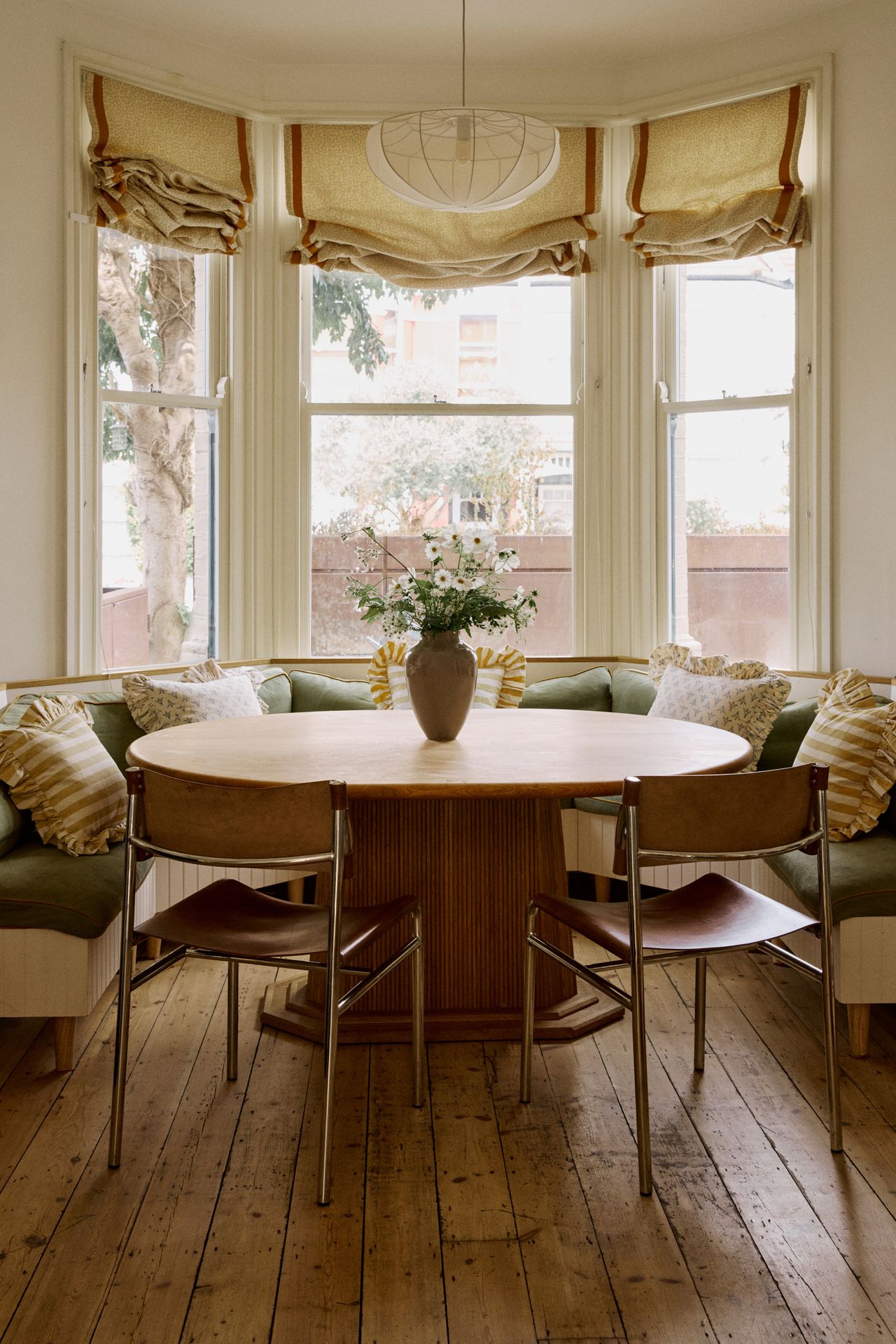 Image of a dining nook in a bay window with a green bench seat that has lots of cushions, a round wooden table, and two leather and metal dining chairs.