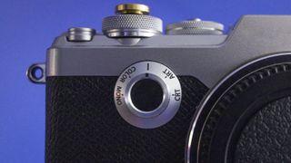 Close-up of an Olympus PEN-F camera body, against a purple background, focusing on the Creative Dial