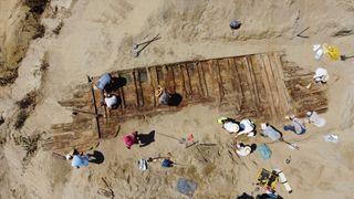 An aerial view of people examining the remains of the wooden boat in the sand.