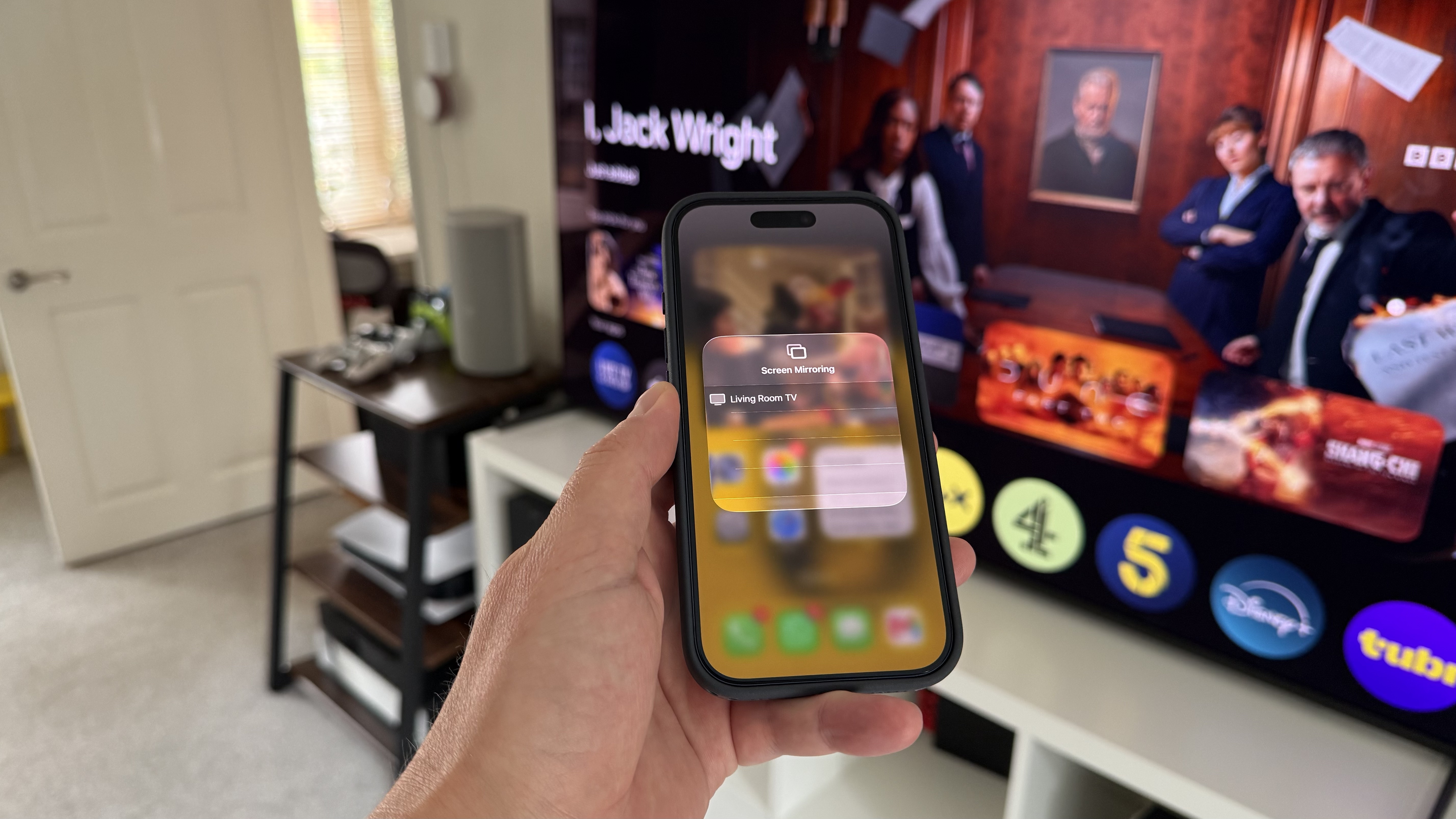 A photo of an iPhone being held in front of a TV. ON the phone screen is the Mirroring menu.