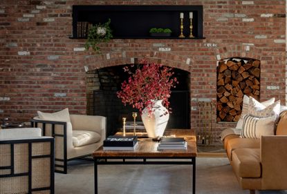 A living room with a brick wall, muted furniture pieces and a coffee table with a dry flower arrangement 