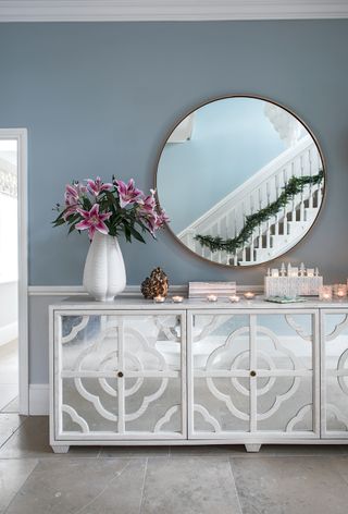 A mirrored sideboard and mirror above in a hallway with light blue walls with a reflection of banisters with a garland