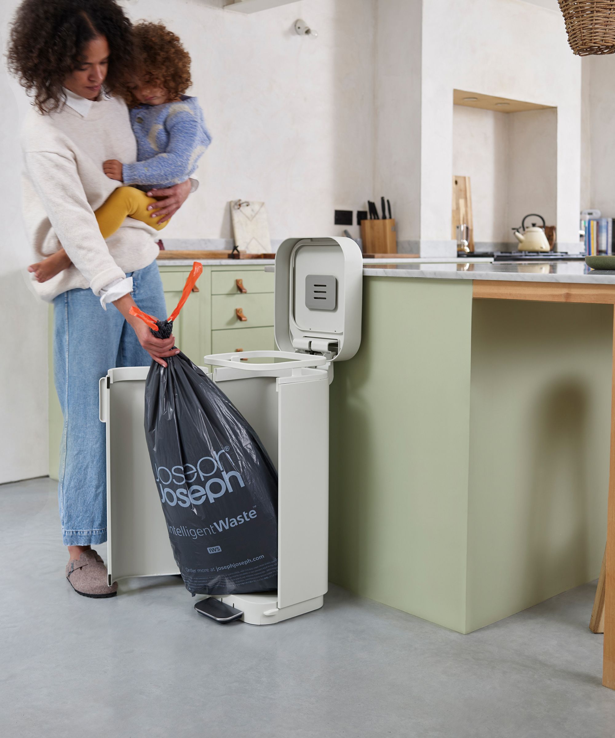A woman holding a child removes a full bin liner from a freestanding white recycling bin with a foot pedal, placed beside a pale green kitchen island.