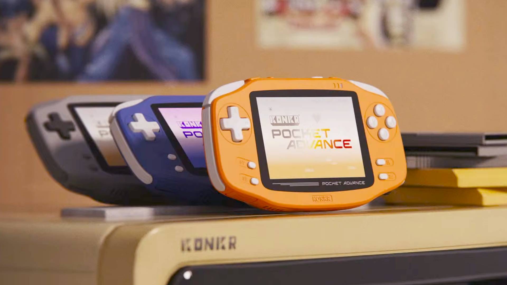 Coral, Navy, and gray KONKR Pocket Advance handhelds in a row on top on faux beige CRT.