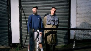 A portrait of Sleaford Mods stood outside a gloomy building