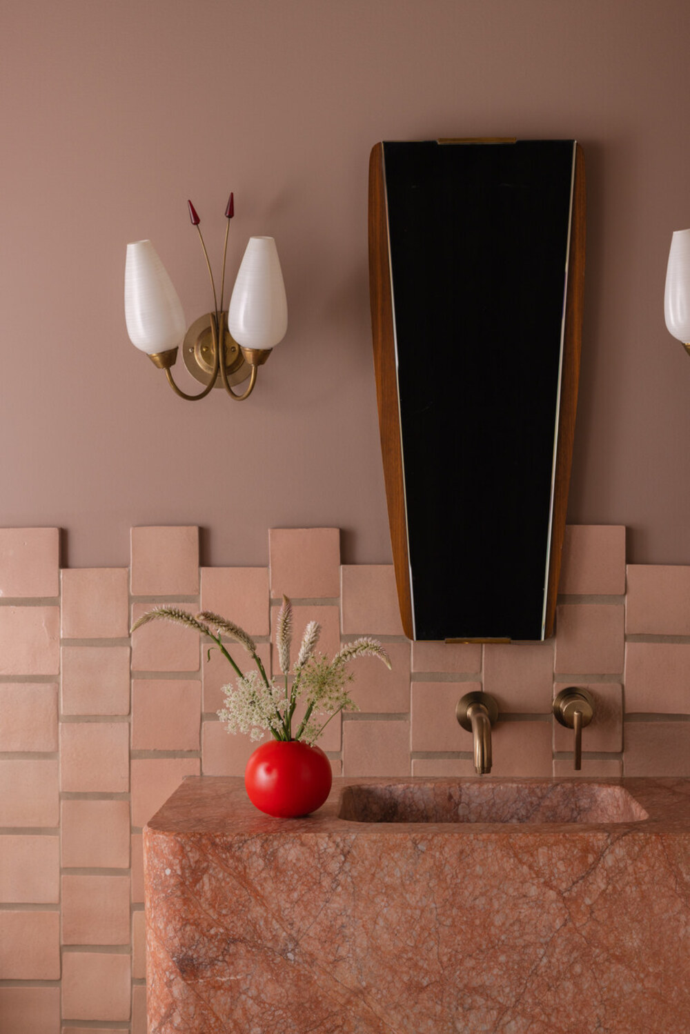 dusty pink bathroom with bricked walls, marble sink, timber mirror, wall sconce, and bright red vase