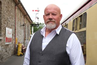 A generic shot of Paul Yellen (Adam Fogerty), bald and bearded, standing on the platform next to the vintage carriages of a steam train