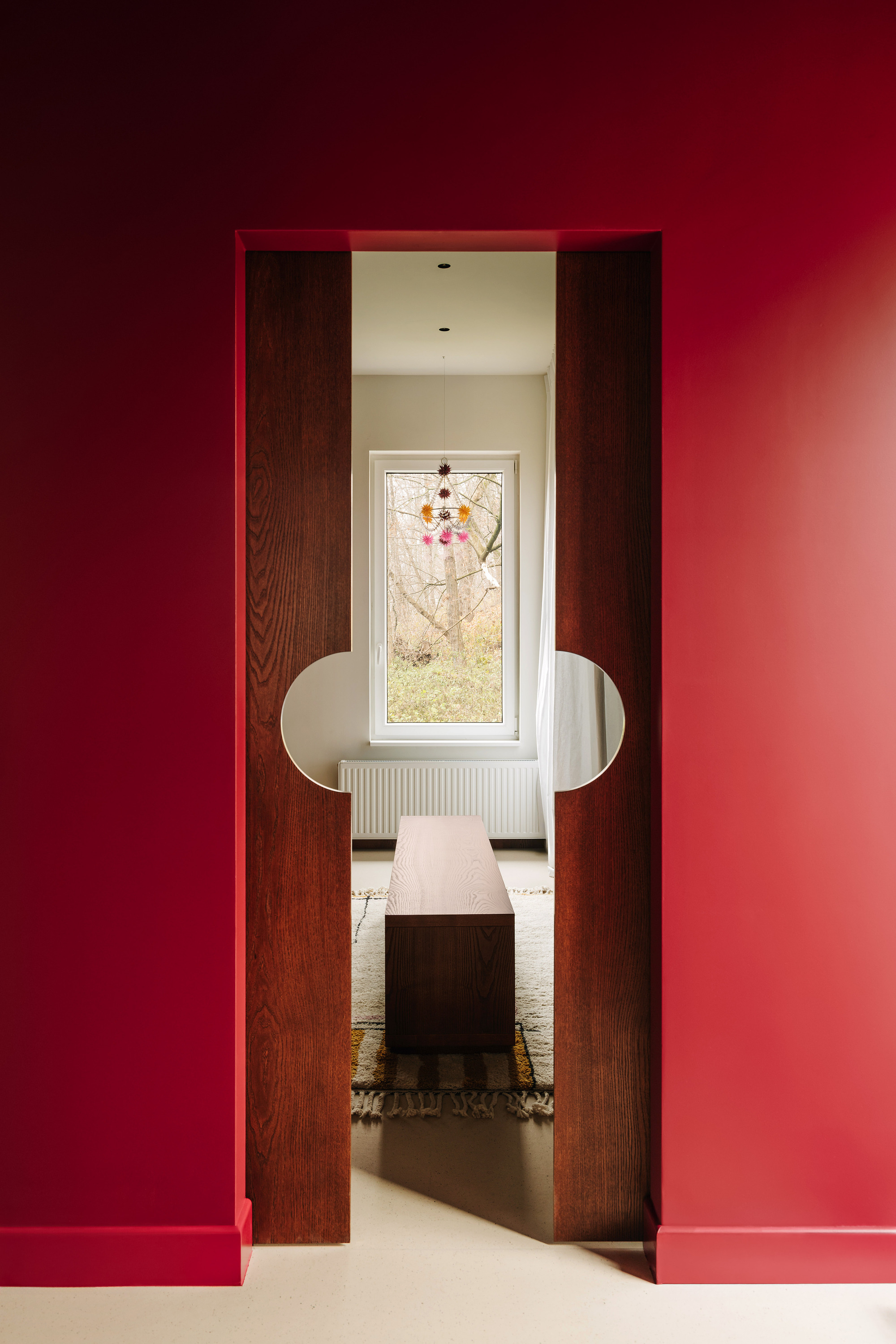 Image of a red wall with a doorway that has wooden sliding doors leading to a white hallway with a rug and wooden bench seat in view.