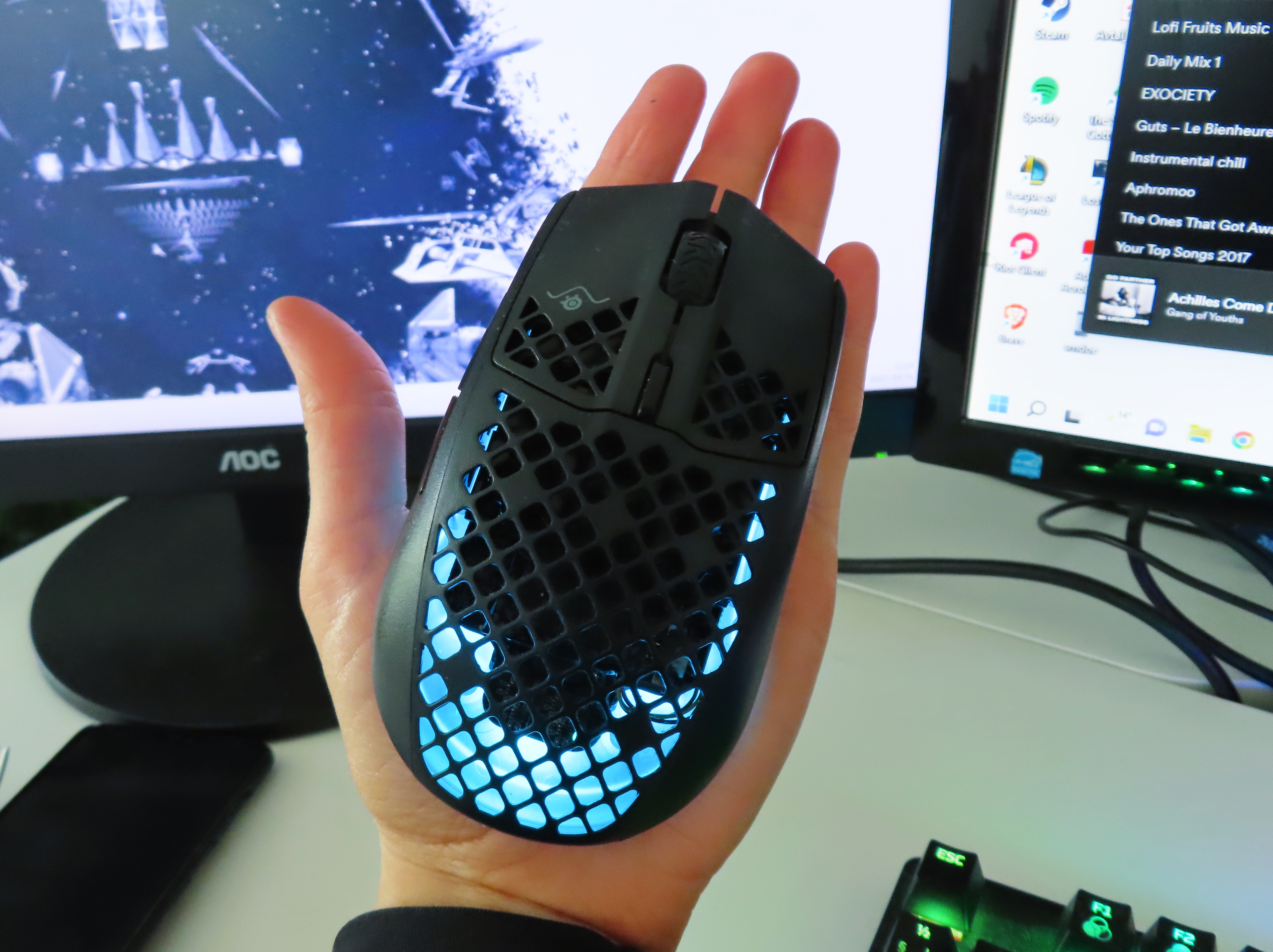 SteelSeries Aerox 3 vilar i en handflata, med en PC-setup i bakgrunden.