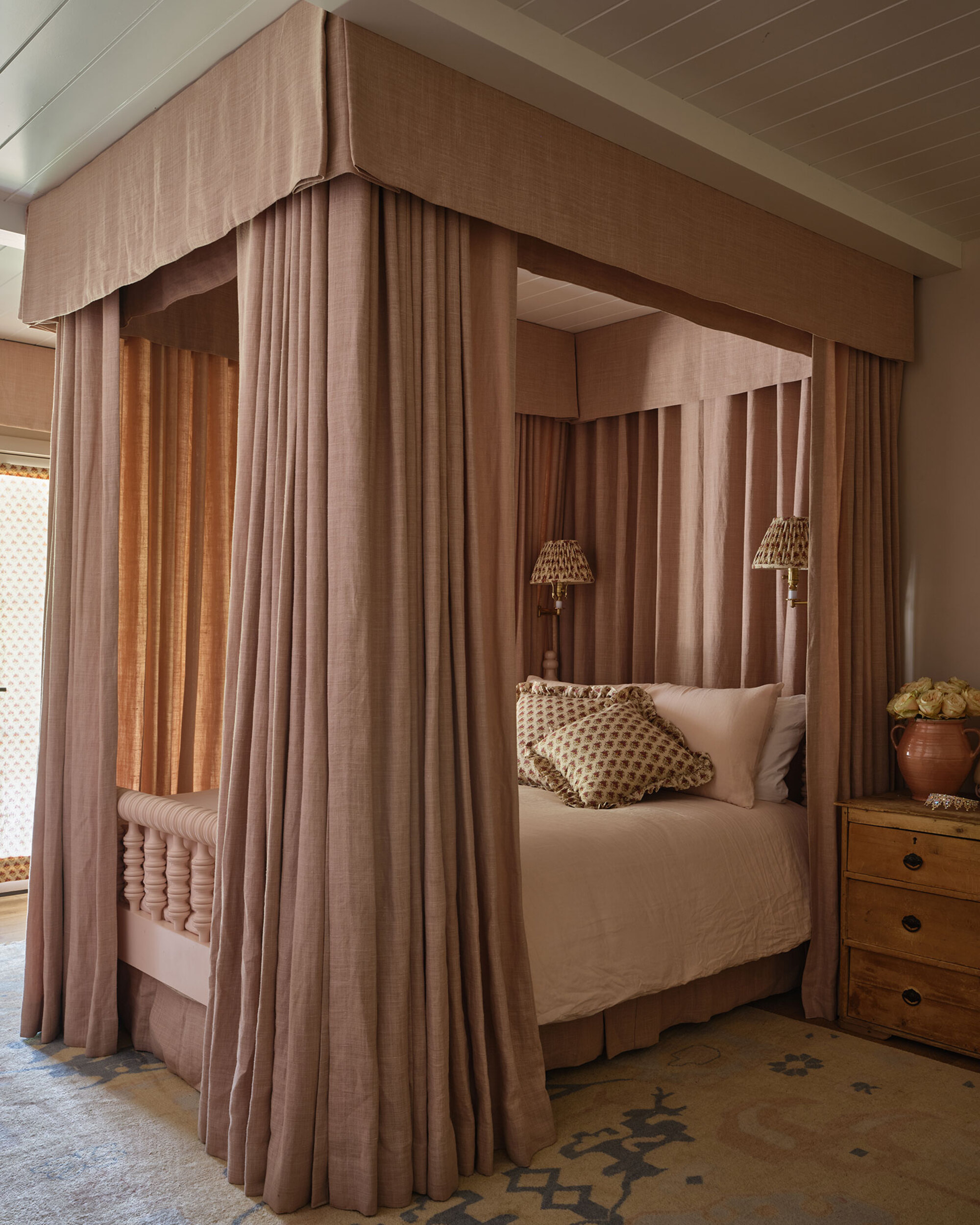 Image of a bed with a dramatic, dusty pink canopy and curtains around it. The bedding is a lighter pink with green patterned curtains.