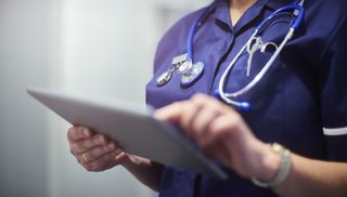 A female surgeon in blue scrubs operating a tablet, to represent the NHS digital experience. She is shot from the neck down, and have a stethoscope around her neck.