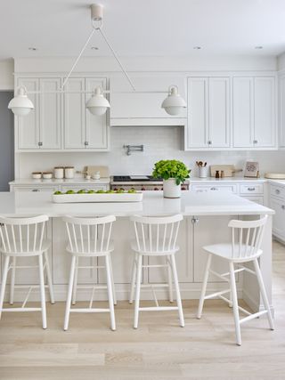 A kitchen with white chairs, counter, backsplash, cabinets and even lighting