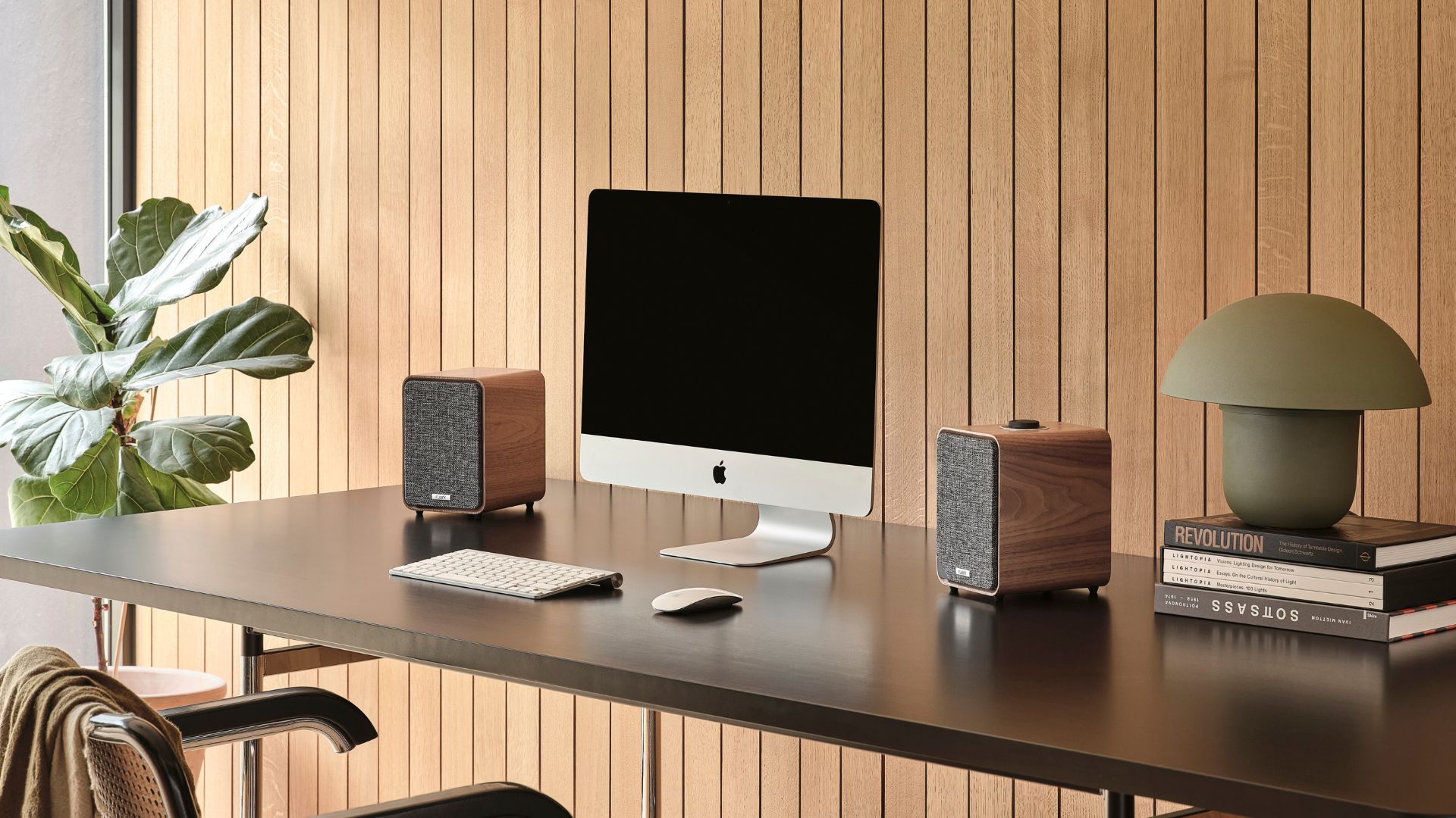 an image of the ruark speakers in walnut on top of a desk with an apple mac on it. The desk is against a wooden slatted wall, with a tall plant next to the desk. 