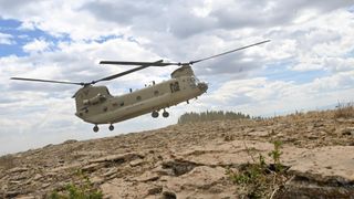 A helicopter with dual propellors floats over the dry, weedy Colorado landscape with a large cloudy sky above.