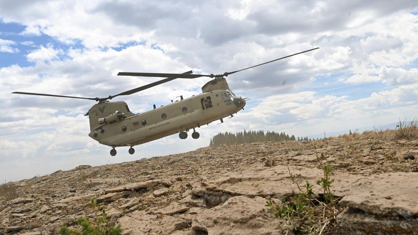 A helicopter with dual propellors floats over the dry, weedy Colorado landscape with a large cloudy sky above.