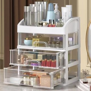 A white and clear plastic makeup organizer with 3 drawers. Cosmetics are exposed inside and above.