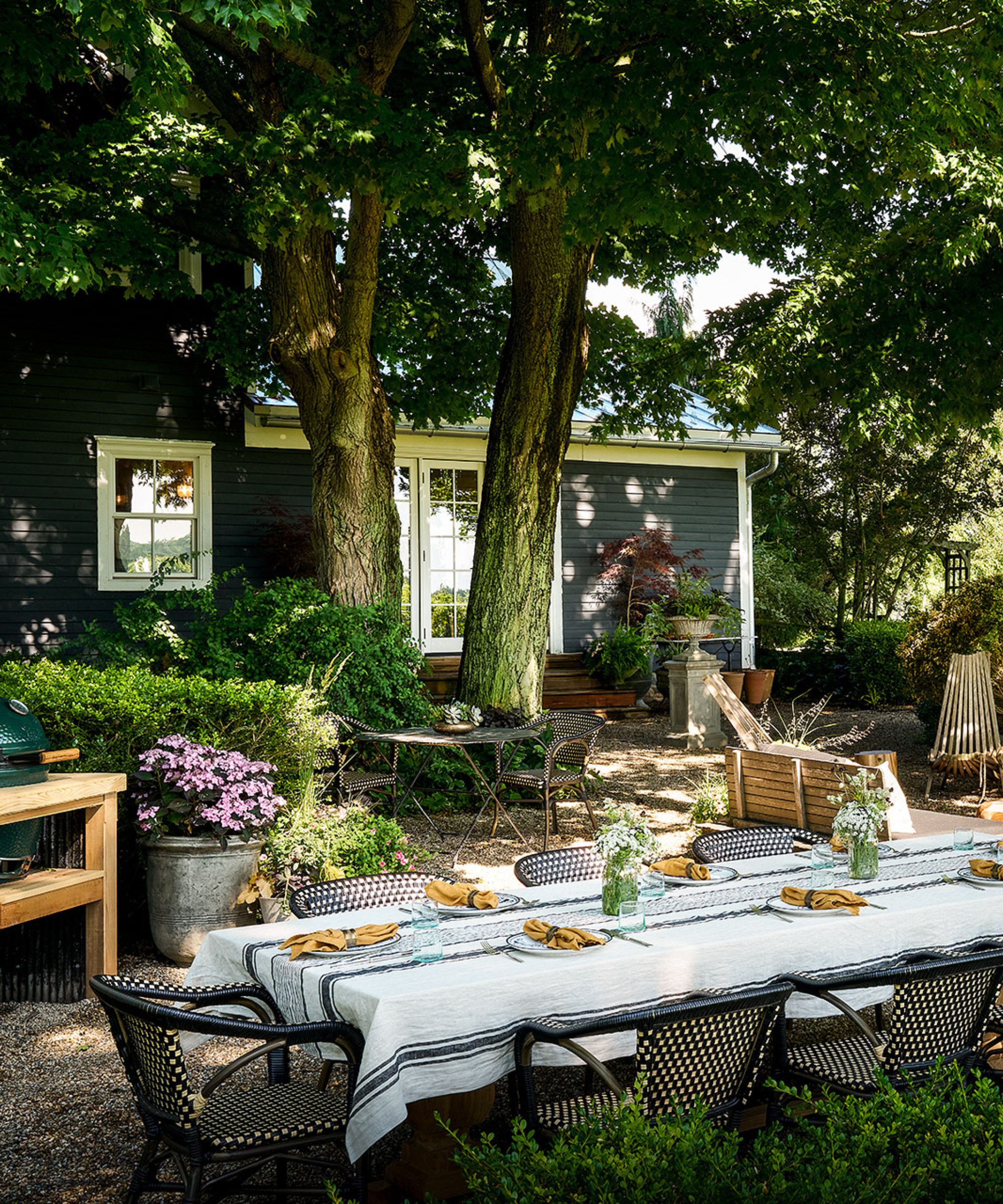 outdoor dining table under the trees with a summer house in the background and a Green Egg barbecue
