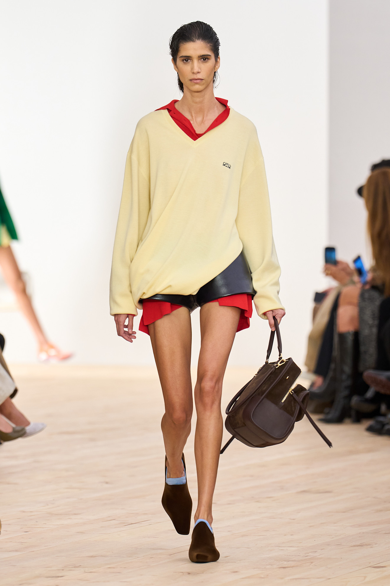 Layering staples for spring 2026 are shown in a photo from Loewe's runway show, with a model walking while wearing a bright red oversized button-down shirt styled under a butter-yellow v-neck sweater and leather shorts, styled with a brown suede handbag and pointed loafers