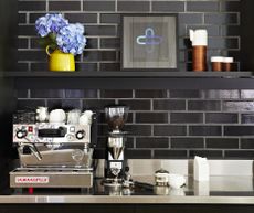 A coffee bar with a black tiled backsplash, a coffee machine, and open shelving displaying flowers in a vase