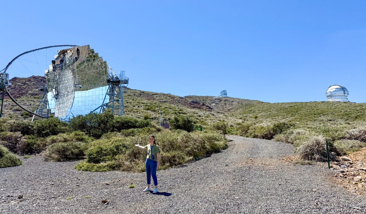 starmus la palma 2025 - a woman stands in the foreground holding her arm out to present a large telescope structure behind her with pristine mirrors reflecting the blue sky above.