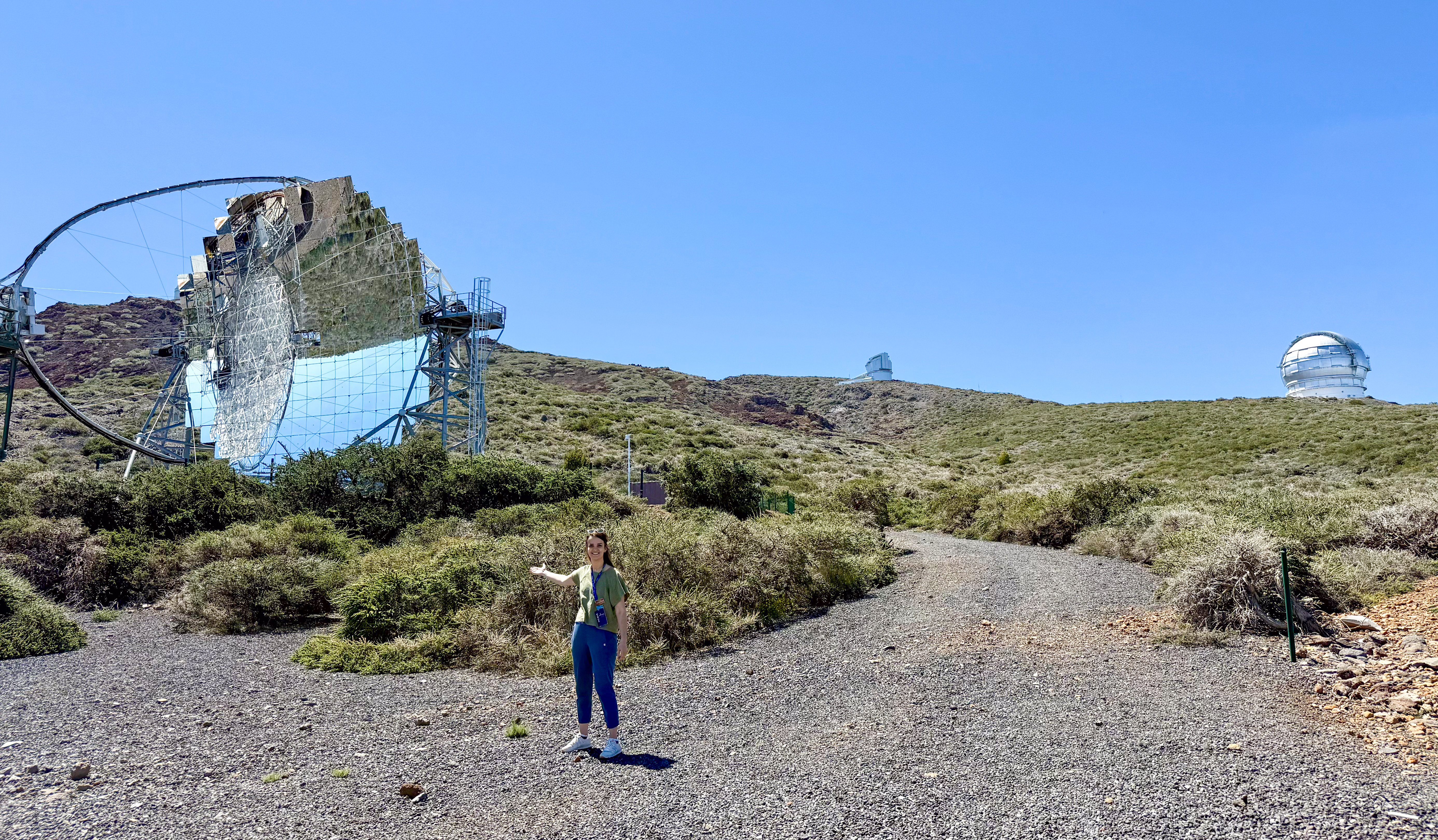 starmus la palma 2025 - a woman stands in the foreground holding her arm out to present a large telescope structure behind her with pristine mirrors reflecting the blue sky above.