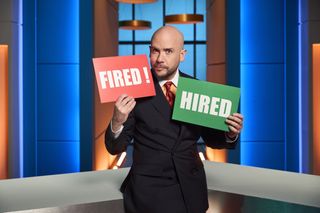 Tom Allen in the brightly lit blue and orange You're Fired studio, wearing a dark suit and colourful tie, holding up a red 'FIRED!' sign and a green 'HIRED!' sign with a raised eyebrow.