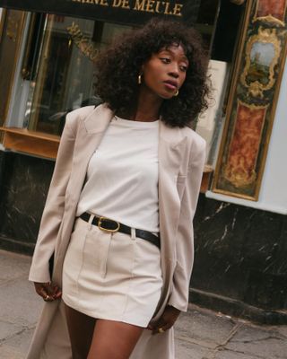 French woman wearing a black belt, a tan miniskirt and jacket, and a white T-shirt