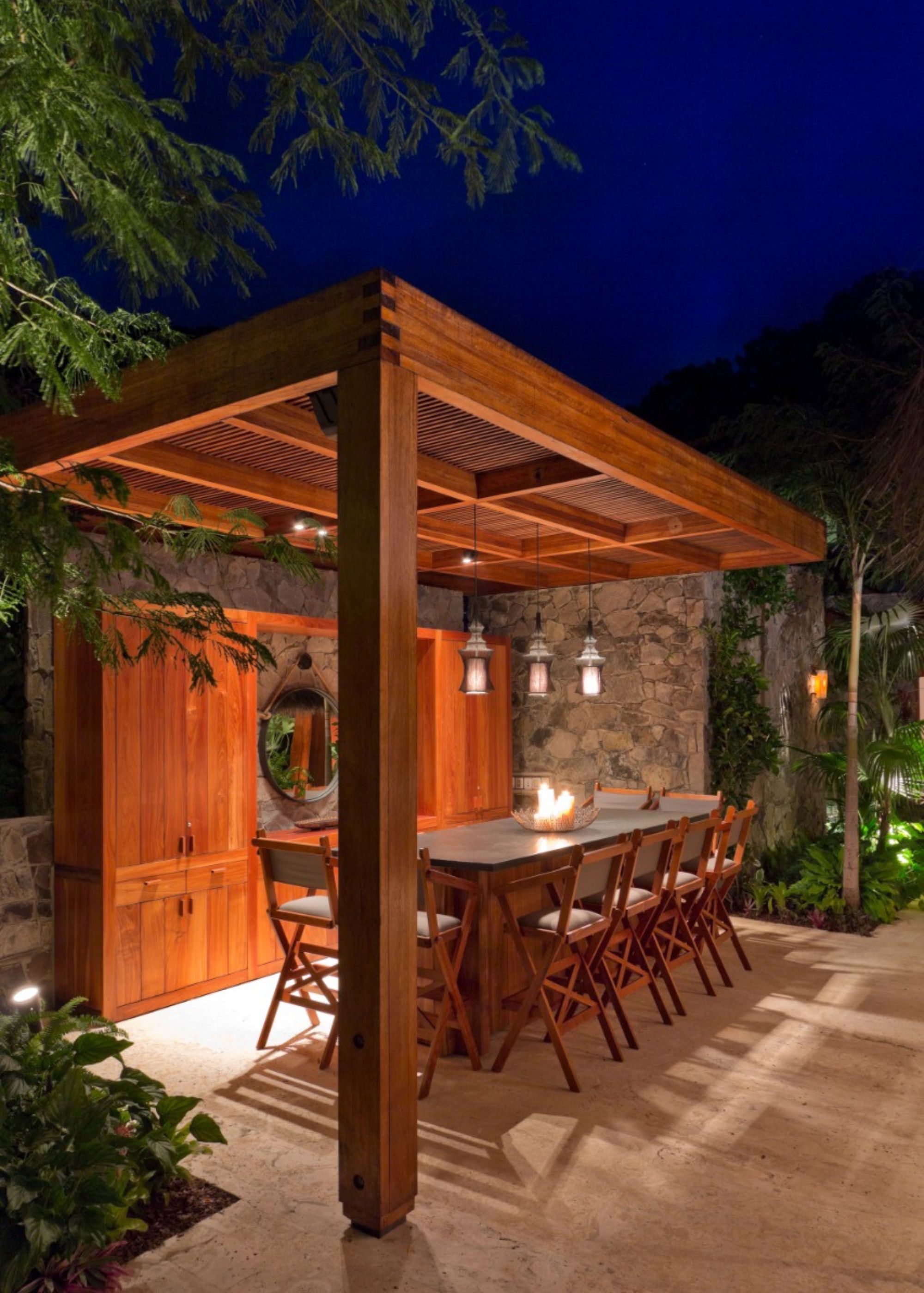 An outdoor dining area with a pergola above it as well as pendant lights hanging from the pergola. Around the dining table are several chairs, and on top of the table is a mini table firepit that has been set alight.