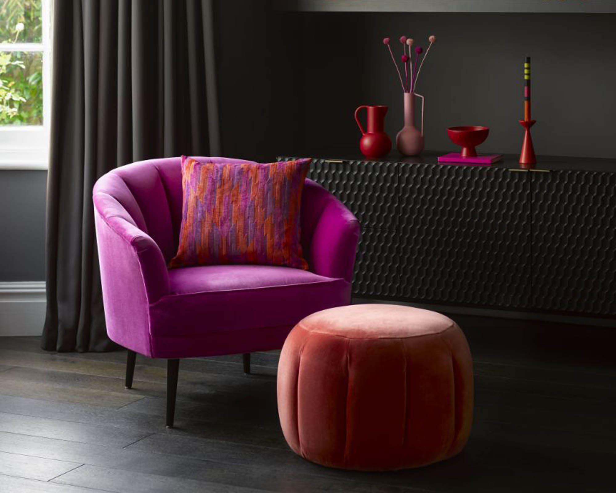 A pink velvet armchair with a curved back, with a cylindrical velvet footstool in orange-red
