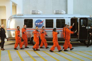 The astronauts are wearing bright orange flight suits and are smiling and waving as they approach the gray astrovan.