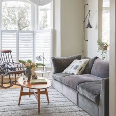 Neutral painted living room with shutters on the window, and a grey sofa on top of a cream rug 