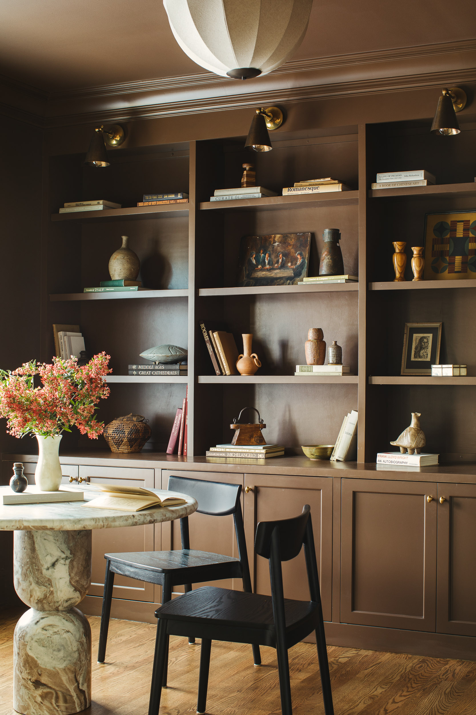 Image of a dark brown bookshelf filled with coffee table books and decorative objects. There is a marble table and two black chairs in front of it.
