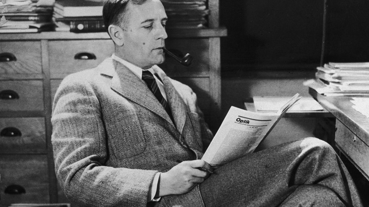 A black and white photo of a man sitting at a desk and reading a magazine.