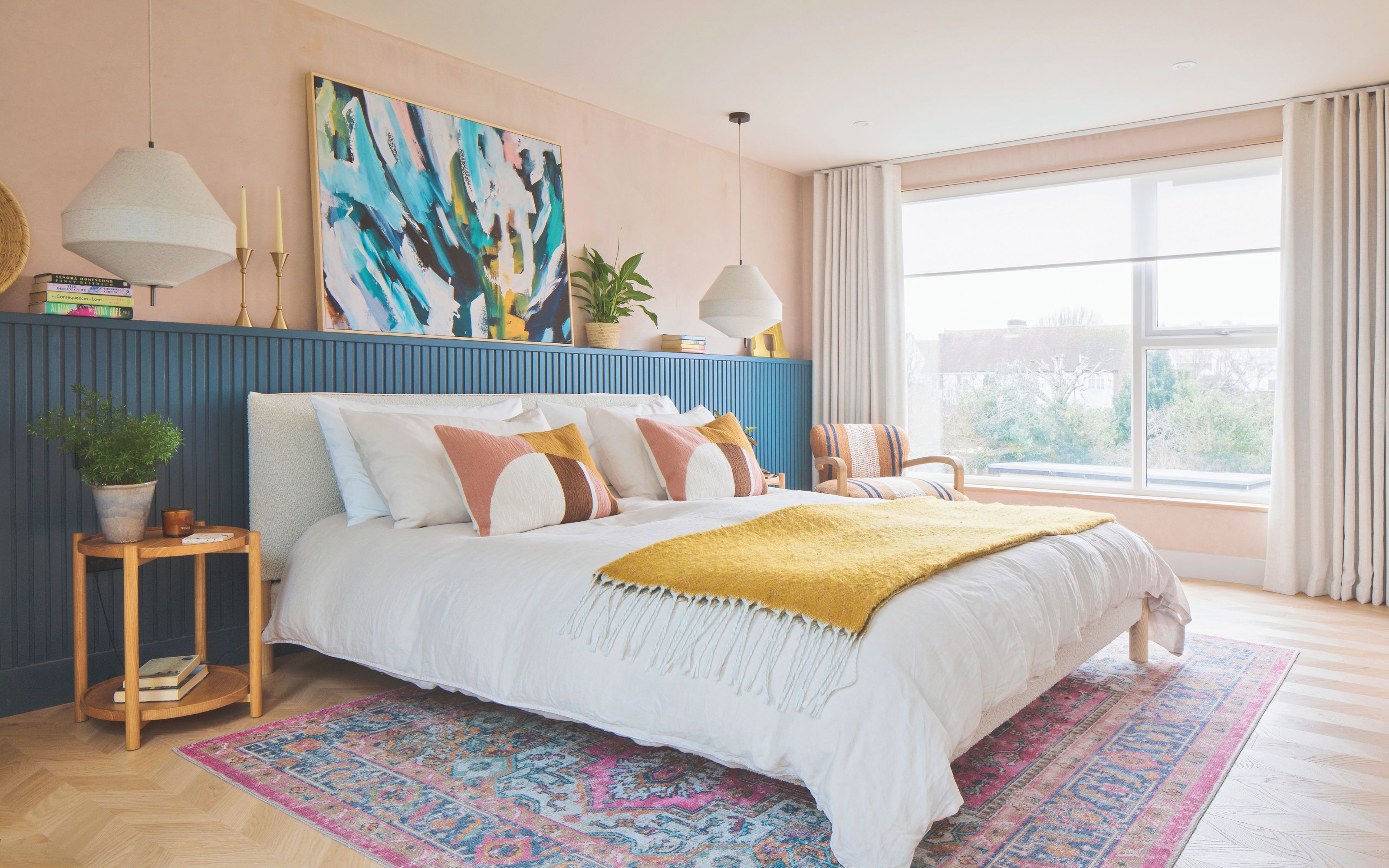 Loft bedroom with large window, pink walls, blue panelling and bed. Pendant lights either side of bed and colourful artwork above with greenery and wooden bedside tables. Mustard blanket on bed.
