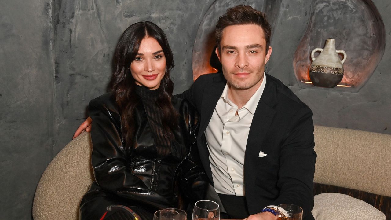 Ed Westwick and Amy Jackson sat on a beige boucle couch with a gray background behind them