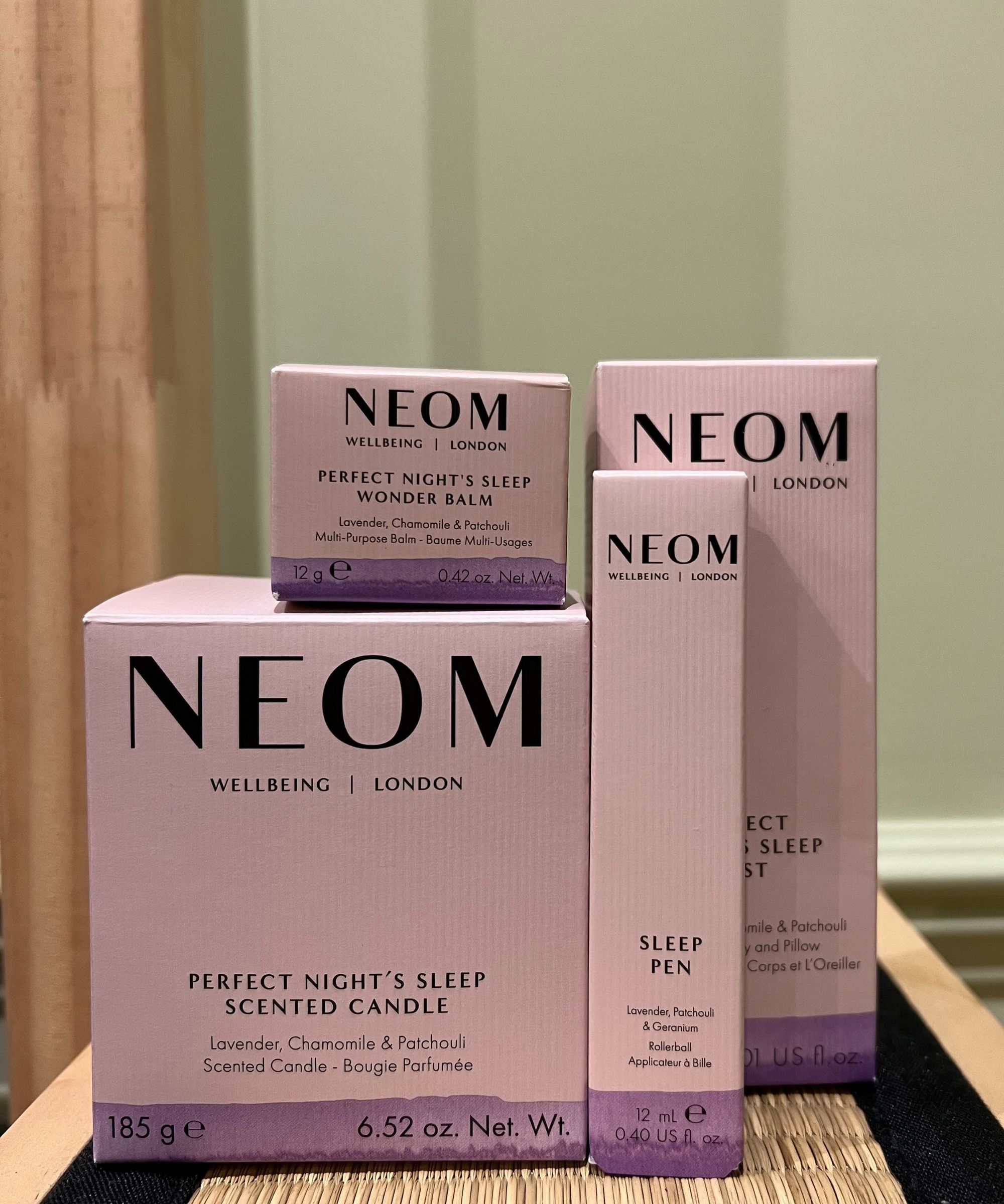 NEOM Perfect Night's Sleep range in pale purple and pink cardboard packaging, on a tatami bedside table, with a green wall behind painted in Farrow &amp;amp; Ball Green Ground.