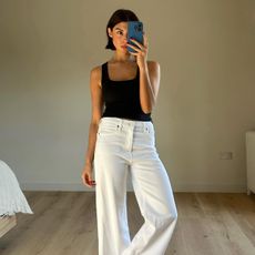 Influencer wearing black tank top and white jeans, taking mirror selfie of full outfit.