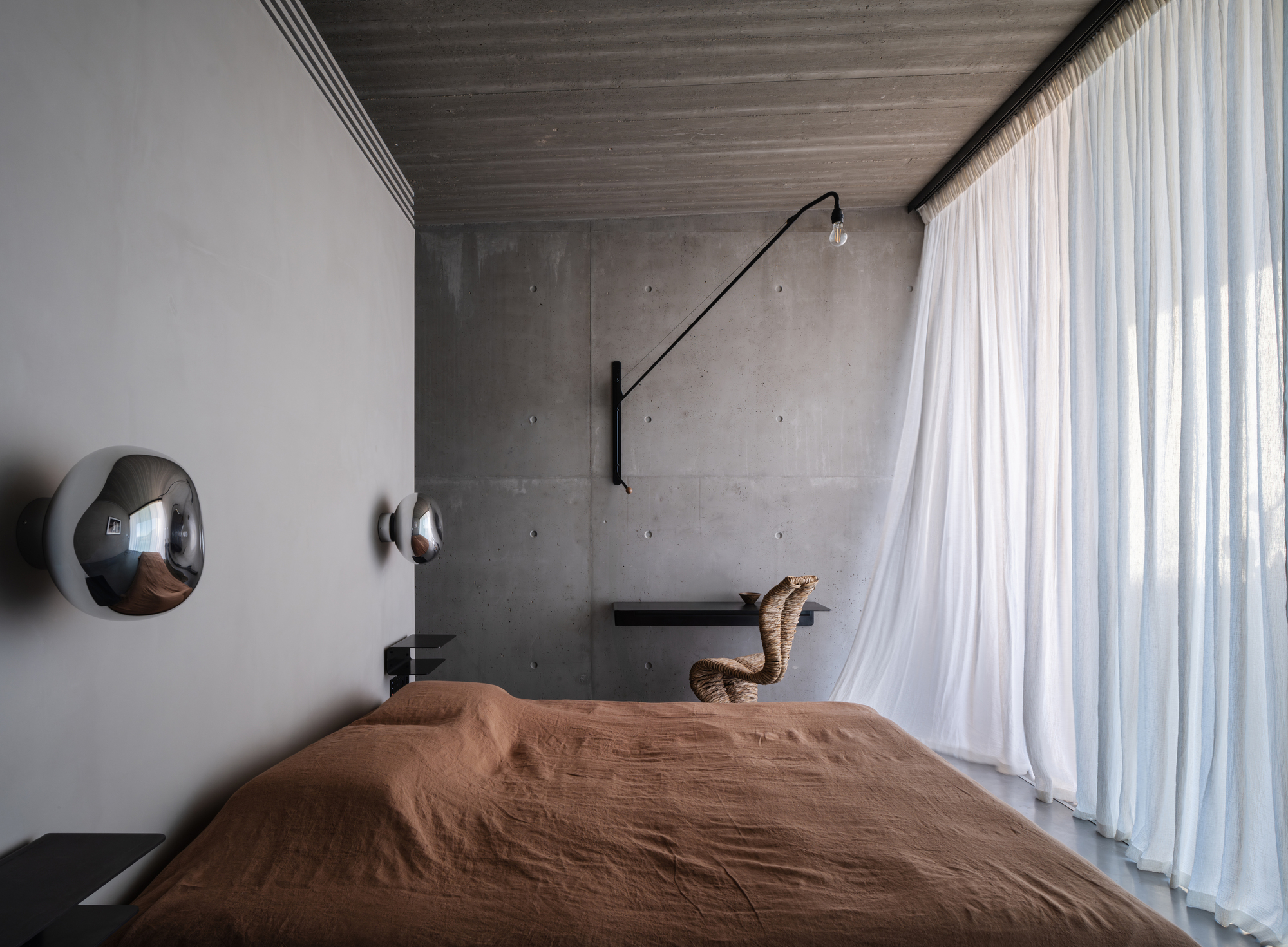 brutalist home bedroom with concrete walls, sheer curtains, orange bedcover on bed, swivel arm lamp