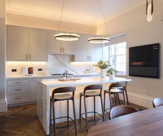 A sophisticated neutral-toned kitchen with a large marble island, complete with waterfall edges. Three dark brown leather barstools line the island, which is set for coffee with a French press and cups. Above, two circular chandeliers with crystal accents add elegance.