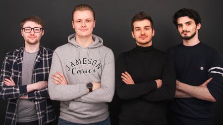 Four young men standing in a line against a dark background, all with their arms crossed, looking at the camera.