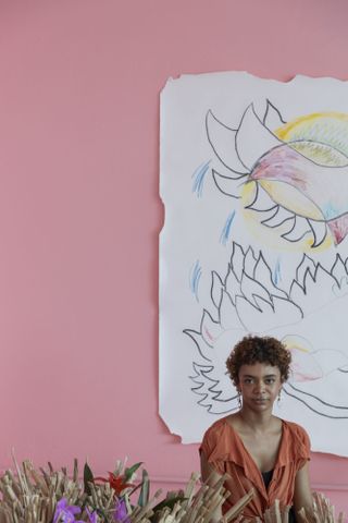 A young woman dressed in an orange blouse sits in front of a large-scale drawing hanging on a pink wall.