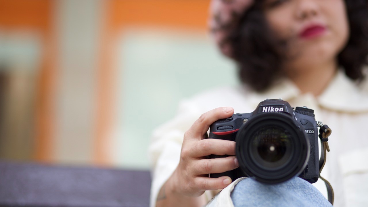 A close-up shot focuses on a person's hands holding a black Nikon D7100 digital SLR camera with a large lens, while the person's face and background are blurred.