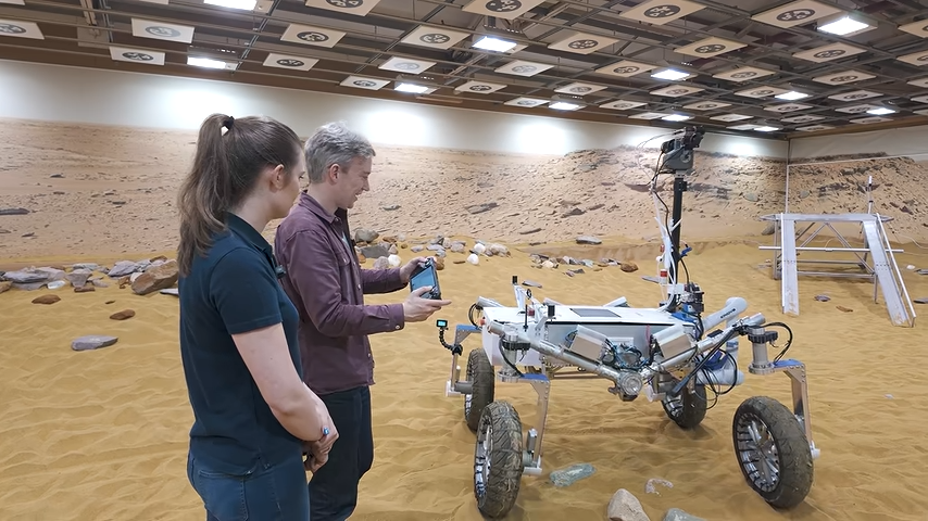 Tom Scott steering a Mars rover prototype with a modified Steam Deck. An Airbus engineer looks on.