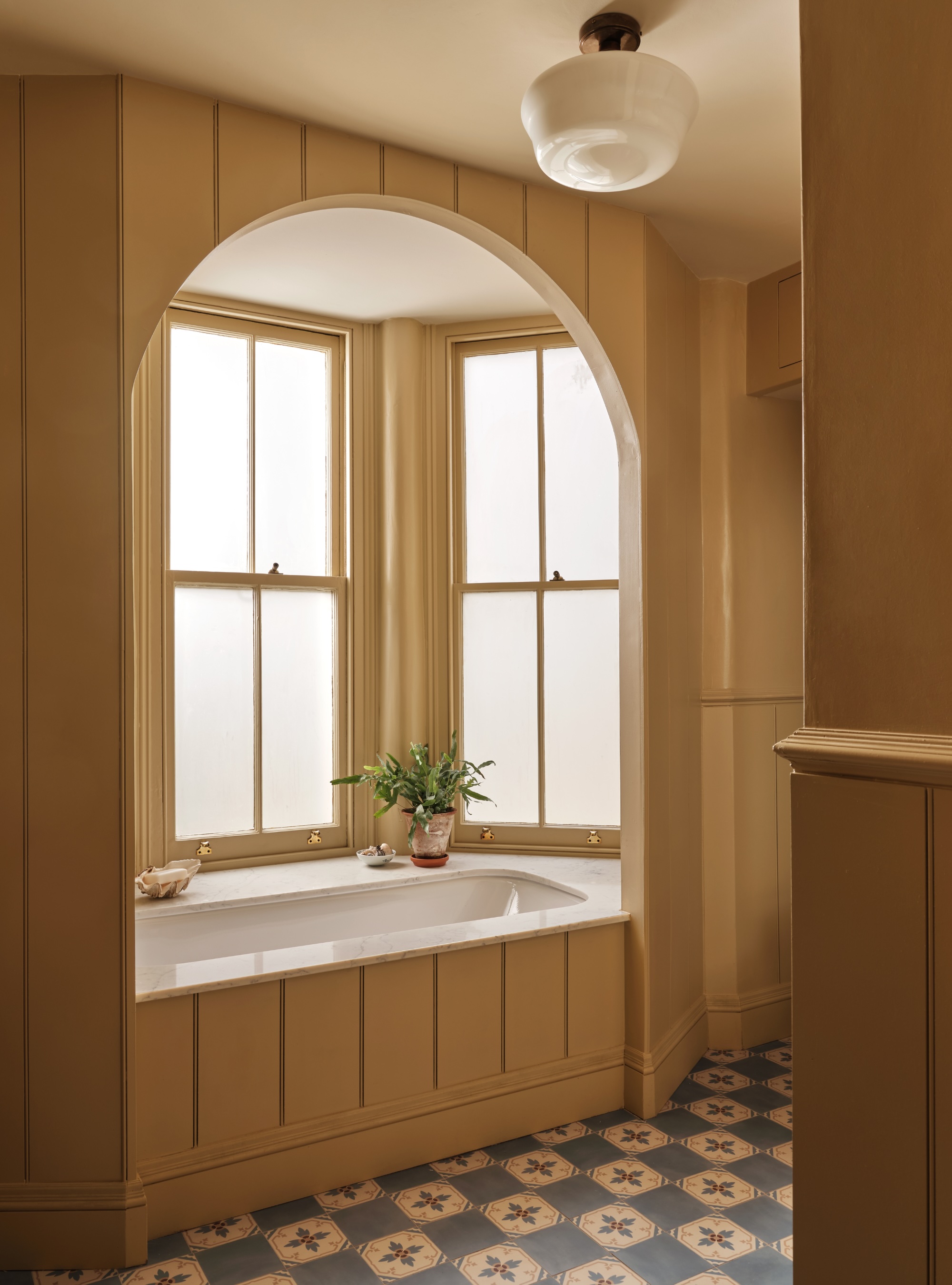 yellow color drenched bathroom with patterned floor, built-in bath in alcove with panelling, frosted windows, plant in pot