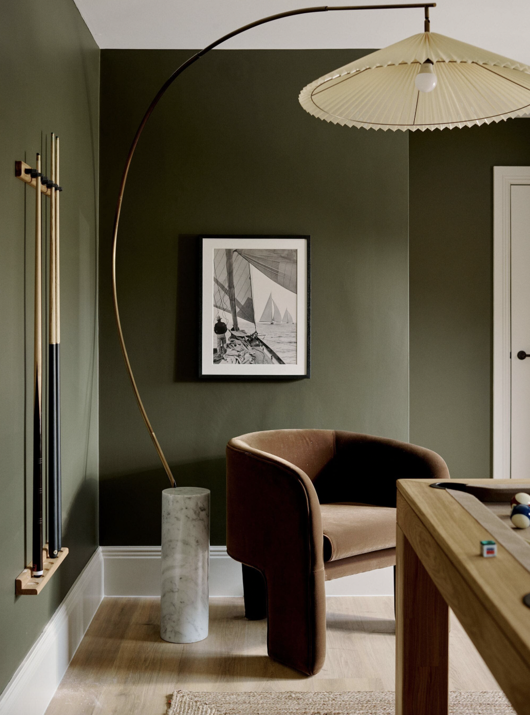 A green room with natural materials with timber floors and a jute rug, a large sculptural lamp, a brown velvet armchair, and a pool table