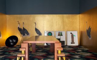 A large dining table is set in the center of the room, on a colorful carpet. The walls are painted in gold with birds on them.