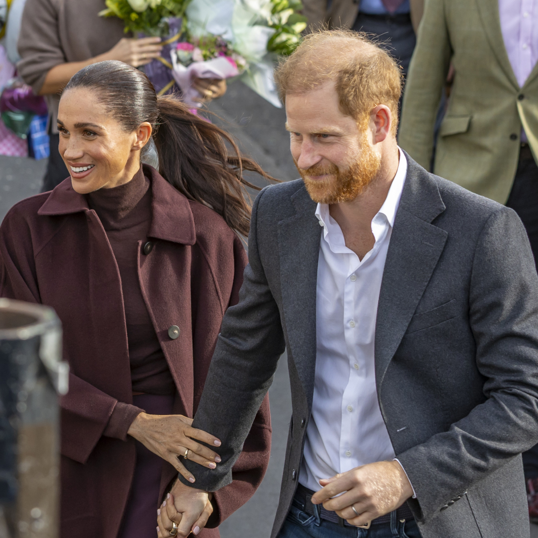 Meghan Markle wearing a maroon coat holding Prince Harry's arm