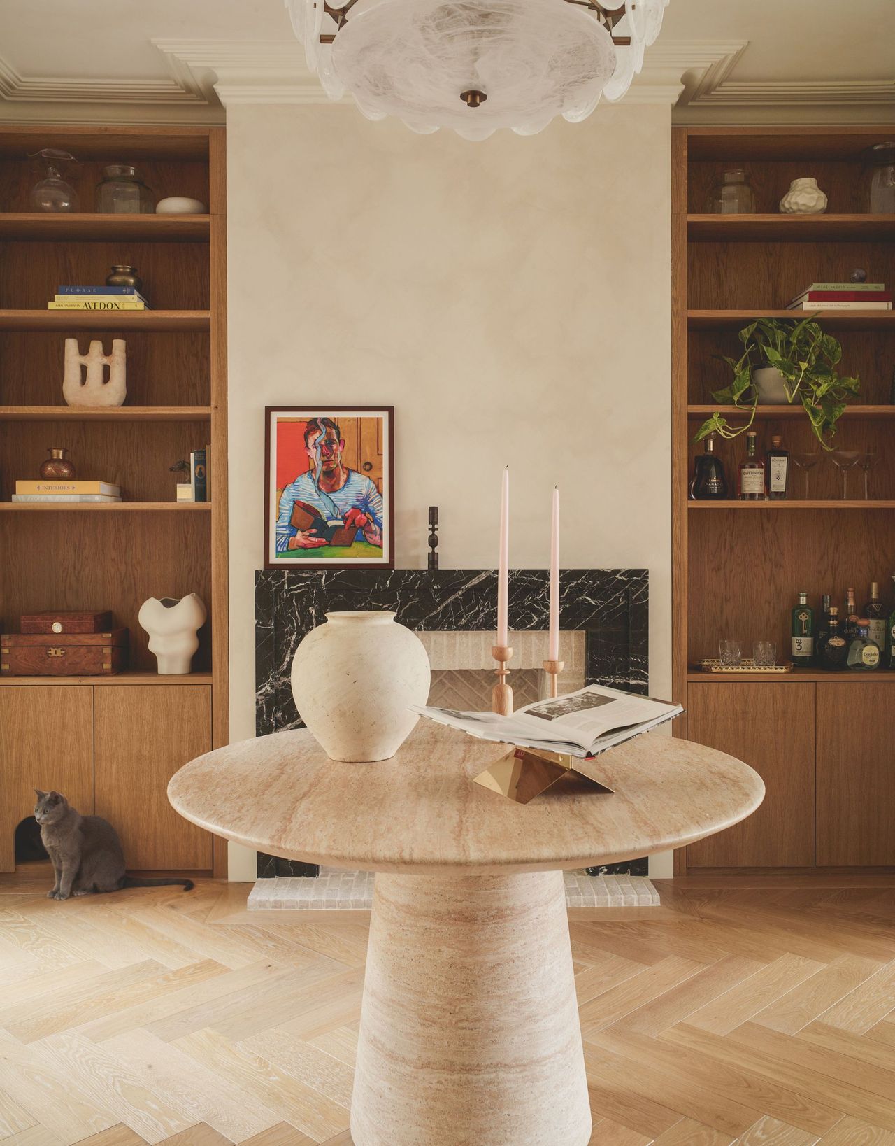 A round stone pedestal table sits in the middle of a room in front of an inset green marble-framed fireplace with alcove shelving either side and herringbone wood flooring