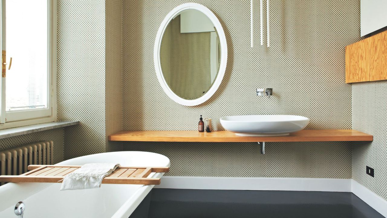 Minimalist bathroom with oval self-lit wall-mounted mirror, floating sink and shelf, bright window and freestanding bath to the left. Neutral dotted wallpaper is set against pops of white and natural wood